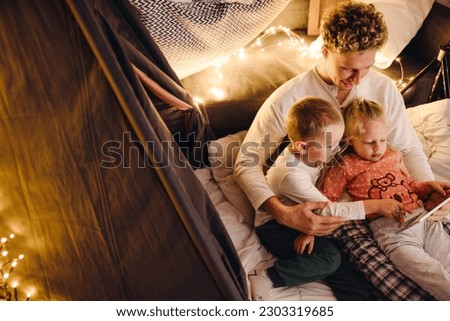 Smiling father and his cute kids wearing pajamas watching cartoons on digital tablet while sitting in handmade tent in children's room