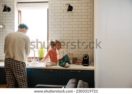 Young father washing dishes while his kids watching cartoons on digital tablet in cozy kitchen