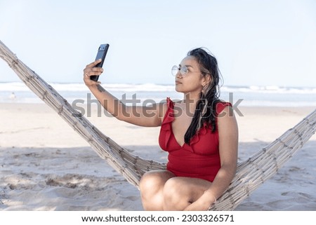A young Hispanic woman is taking a selfie photo on a hammock at the beach