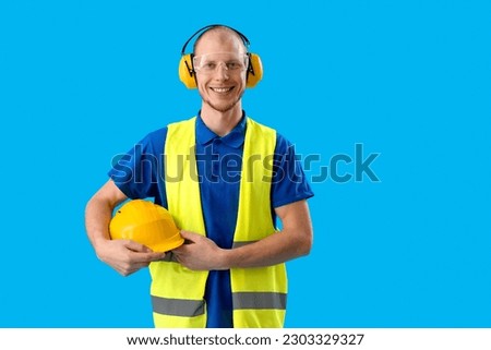 Male worker in vest, with hardhat and protective headphones on blue background Royalty-Free Stock Photo #2303329327