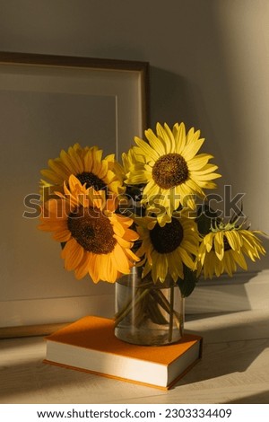 A bouquet of sunflowers in a glass vase, a hardcover book, a poster frame against the wall in the warm sunset light.
