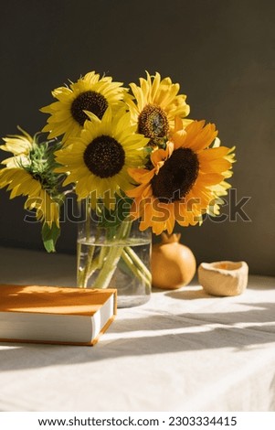 A bouquet of sunflowers in a glass vase, a hardcover book, ceramics on linen tablecloth in the warm sunlight. Still life.