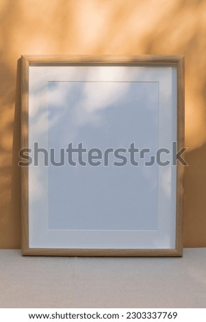 Blank wooden frame mockup against beige wall with dappled light coming through the leaves. A showcase of works of art. 