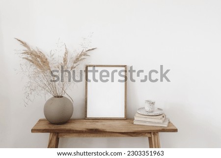 Ball shaped vase with dry grass bouquet. Old wooden bench. Blank vertical picture frame mockup with books and cup of coffee, tea. White wall background. Empty copy space. Neutral still life interior.