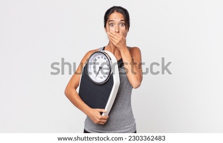young hispanic woman covering mouth with hands with a shocked and holding a weight scale. diet concept Royalty-Free Stock Photo #2303362483