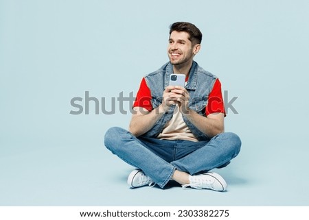 Full body young man he wear denim vest red t-shirt casual clothes hold in hand use mobile cell phone look aside on area isolated on plain pastel light blue cyan background studio. Lifestyle concept