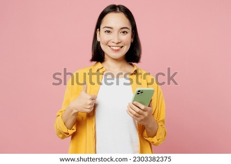Young fun woman of Asian ethnicity wear yellow shirt white t-shirt hold in hand use mobile cell phone show thumb up isolated on plain pastel light pink background studio portrait. Lifestyle concept