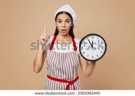 Young housewife housekeeper chef baker latin woman wear striped apron toque hat hold in hands clock point finger up warn about time isolated on plain pastel light beige background. Cook food concept Royalty-Free Stock Photo #2303382445