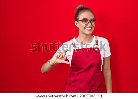 Young hispanic woman wearing waitress apron over red background smiling and confident gesturing with hand doing small size sign with fingers looking and the camera. measure concept.  Royalty-Free Stock Photo #2303386111