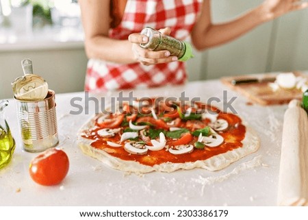 Young beautiful hispanic woman pouring oregano on pizza at the kitchen