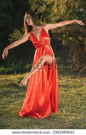 beautiful woman in an elegant red evening dress on the green grass of a beautiful natural landscape at sunset, with her arms poised in balance and showing off her toned dancer's leg out of the dress. 