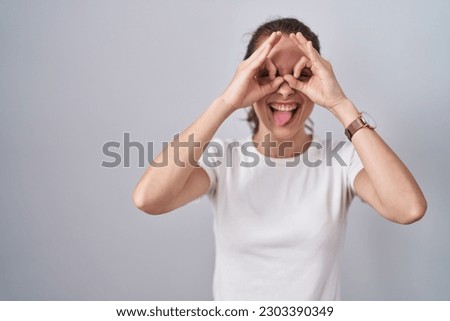Beautiful brunette woman standing over isolated background doing ok gesture like binoculars sticking tongue out, eyes looking through fingers. crazy expression. 