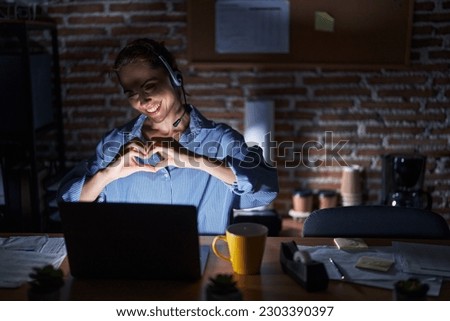 Beautiful brunette woman working at the office at night smiling in love doing heart symbol shape with hands. romantic concept. 