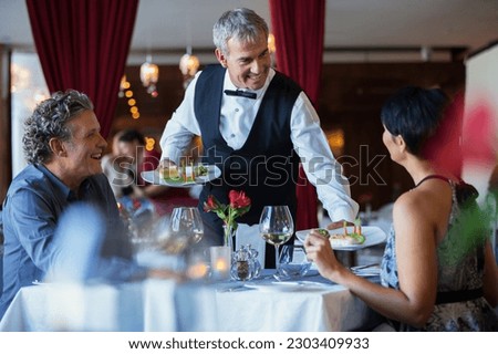 Smiling waiter serving fancy dishes to mature couple sitting at table in restaurant Royalty-Free Stock Photo #2303409933