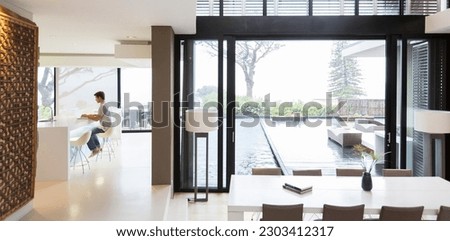 Young man using laptop in modern kitchen, dining area patio door swimming pool in foreground