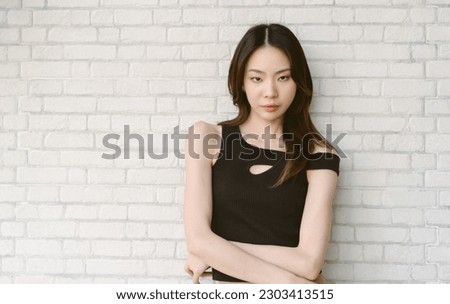 Calm serious beautiful Asian woman wearing off shoulder black and posing with crossed arms and looking at camera on white brick wall background. Attractive young adult gen y lady face without smile.