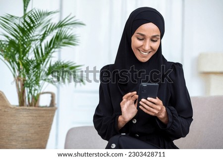 Young Muslim student woman in black hijab sitting at home in hotel and smiling using mobile phone. Corresponds, reads news, checks social networks.