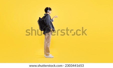 Side View Of Young Man With Backpack Wearing Headphones Listening To Music And Pressing Invisible Button Or Showing Free Space Standing On Yellow Studio Background. Panorama