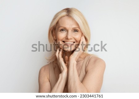 Portrait of happy good-looking senior woman with beautiful skin smiling at camera and touching flawless face skin, posing on white wall background Royalty-Free Stock Photo #2303441761