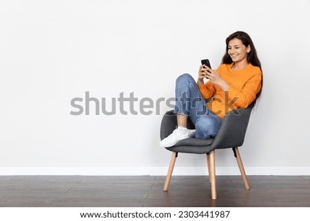 Relaxed happy beautiful young indian woman chilling in armchair over empty white wall background, using cell phone and smiling, scrolling on social media, chatting with friends, copy space
