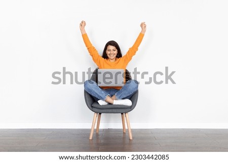 Emotional joyful pretty young indian woman wearing casual outfit sitting in armchair over white background, using laptop, raising hands up, celebrating success, trading on stocks, copy space