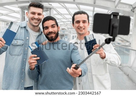 Travelers Selfie. Three Joyful Tourists Men Posing With Passports And Boarding Passes Taking Photo On Smartphone, Having Fun Before Flight In Modern Airport. Share Your Vacation Travel Moments