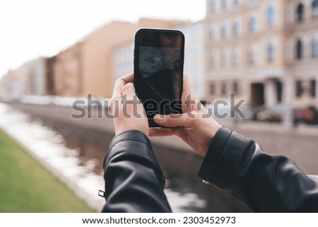 Crop anonymous person in black trendy outfit taking photo on smartphone while standing on blurred background of city street during summer trip