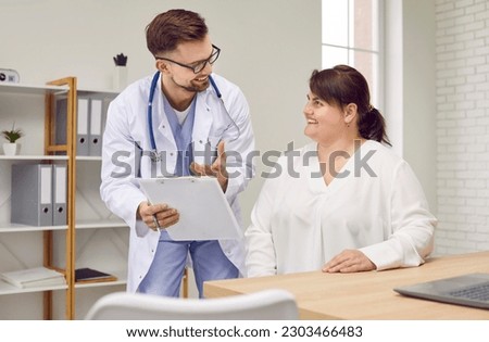 Overweight woman having consultation at doctor's office. Portrait of smiling doctor holding report file with appointment and giving consultation to a fat patient during medical examination in clinic. Royalty-Free Stock Photo #2303466483