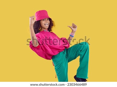 Little kid in trendy clothes. Happy child having fun in fashion studio. Afro American girl wearing fuschia bucket hat, loose oversized top and green jeans dancing isolated on solid yellow background