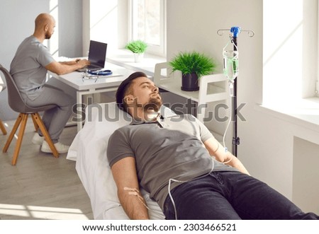 Male patient receiving intravenous therapy treatment at clinic. Young man getting vitamin or medication solution through IV line infusion while lying on medical bed in room with nurse in background Royalty-Free Stock Photo #2303466521