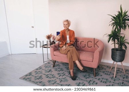 Full body of senior woman in smart casual sitting on sofa near green plant and coffee table and using smartphone for taking photo smiling