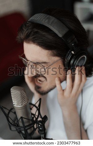 Side view of passionate male singer in headphones sitting at table near microphone and recording song in modern studio