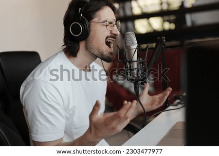 Side view of passionate male singer in headphones sitting at table near microphone and recording song in modern studio