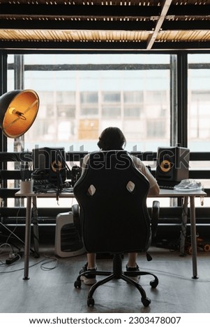 Back view of unrecognizable male musician sitting at table with loudspeaker enclosure near panoramic window and composing music Royalty-Free Stock Photo #2303478007