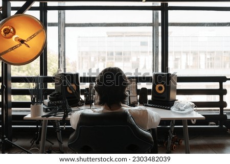 Back view of unrecognizable male musician sitting at table with loudspeaker enclosure near panoramic window and composing music Royalty-Free Stock Photo #2303482039