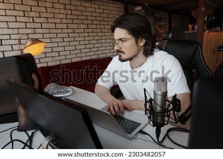 Smiling bearded male musician in eyeglasses and white t shirt composing music on laptop while recording podcast in studio with modern equipment