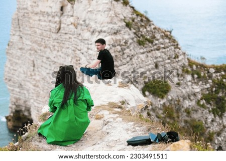 One young caucasian couple of tourists from the back sits high in the mountains and takes pictures on a smartphone near the north sea in Etretat, Normandy, France, close-up view from below.