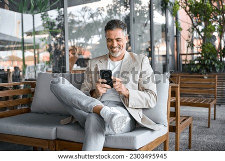 Happy smiling relaxed mid aged business man, mature professional businessman entrepreneur sitting in outdoor cafe holding smartphone using mobile phone digital technology apps. Authentic shot