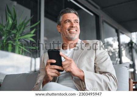 Happy smiling confident mid aged business man, mature professional businessman sitting in outdoor office holding smartphone using mobile phone digital tech looking away thinking. Authentic shot Royalty-Free Stock Photo #2303495947