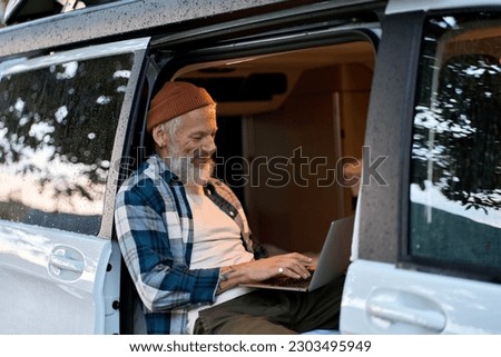 Happy older man sitting in rv camper van using laptop. Smiling mature active traveler holding computer on lap remote working online and enjoying vanlife, freedom, resting in outdoor camping. Royalty-Free Stock Photo #2303495949