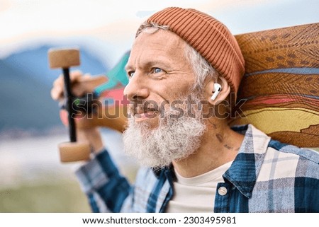 Active cool bearded old hipster man standing in nature park holding skateboard wearing earbud. Mature traveler skater enjoying freedom spirit and extreme sports hobby listening music in earphones.