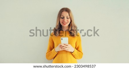 Portrait of happy smiling young woman looking at smartphone wearing knitted yellow sweater on gray background