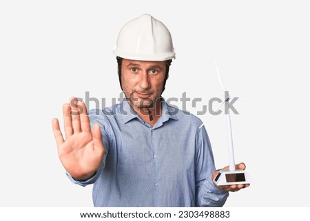 A middle-aged architect man holding a windmill model standing with outstretched hand showing stop sign, preventing you.