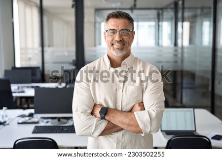 Handsome hispanic senior business man with crossed arms smiling at camera. Indian or latin confident mature good looking middle age leader male businessman on blur office background with copy space.  Royalty-Free Stock Photo #2303502255