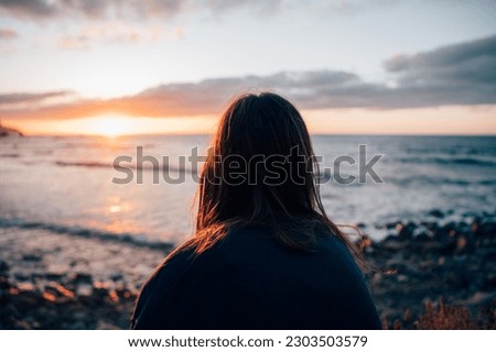 Young girl from behind looking at the sunset at the beach Royalty-Free Stock Photo #2303503579