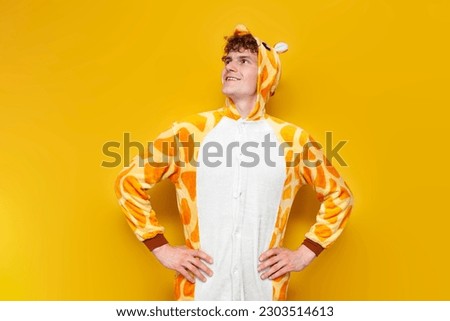 young joyful guy in funny baby giraffe pajamas looks away at the copy space on yellow background, man in animal cosplay clothes, pajama party concept