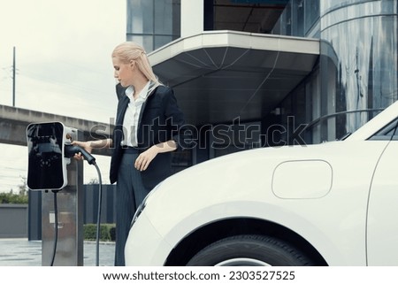 Progressive businesswoman insert charger plug from charging station to her electric vehicle with apartment condo building in background. Eco friendly rechargeable car powered by sustainable energy.