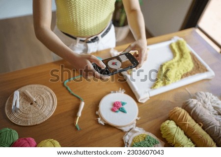 Asian Woman taking smartphone photo Punch needle. phone posting on social networks in studio workshop. designer workplace Handmade craft project DIY embroidery.