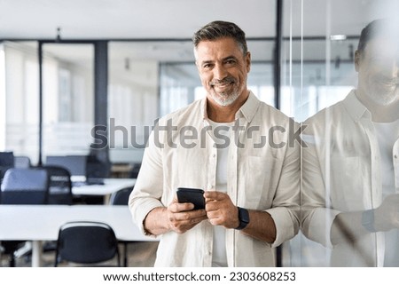 Smiling mature Latin or Indian businessman holding smartphone, using cellphone mobile app. Middle aged hispanic employee standing with phone and looking at camera on office background with copy space.