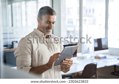 Happy Latin Hispanic bearded stylish mature adult professional business man, smiling Indian senior businessman CEO holding digital tablet using fintech tab application standing inside company office. Royalty-Free Stock Photo #2303623833